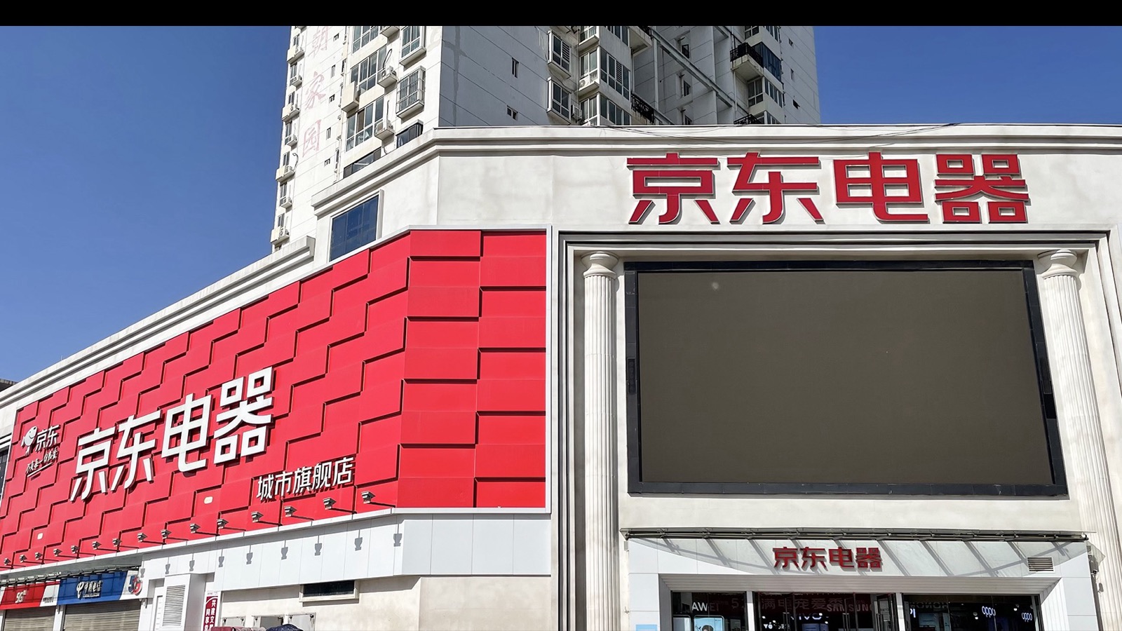 河南省商丘市の「京東電器」都市旗艦店。JD.com が自社ブランドで展開する家電量販店で、正面と側面に赤い京東ロゴとマスコット犬 Joy が並ぶ。