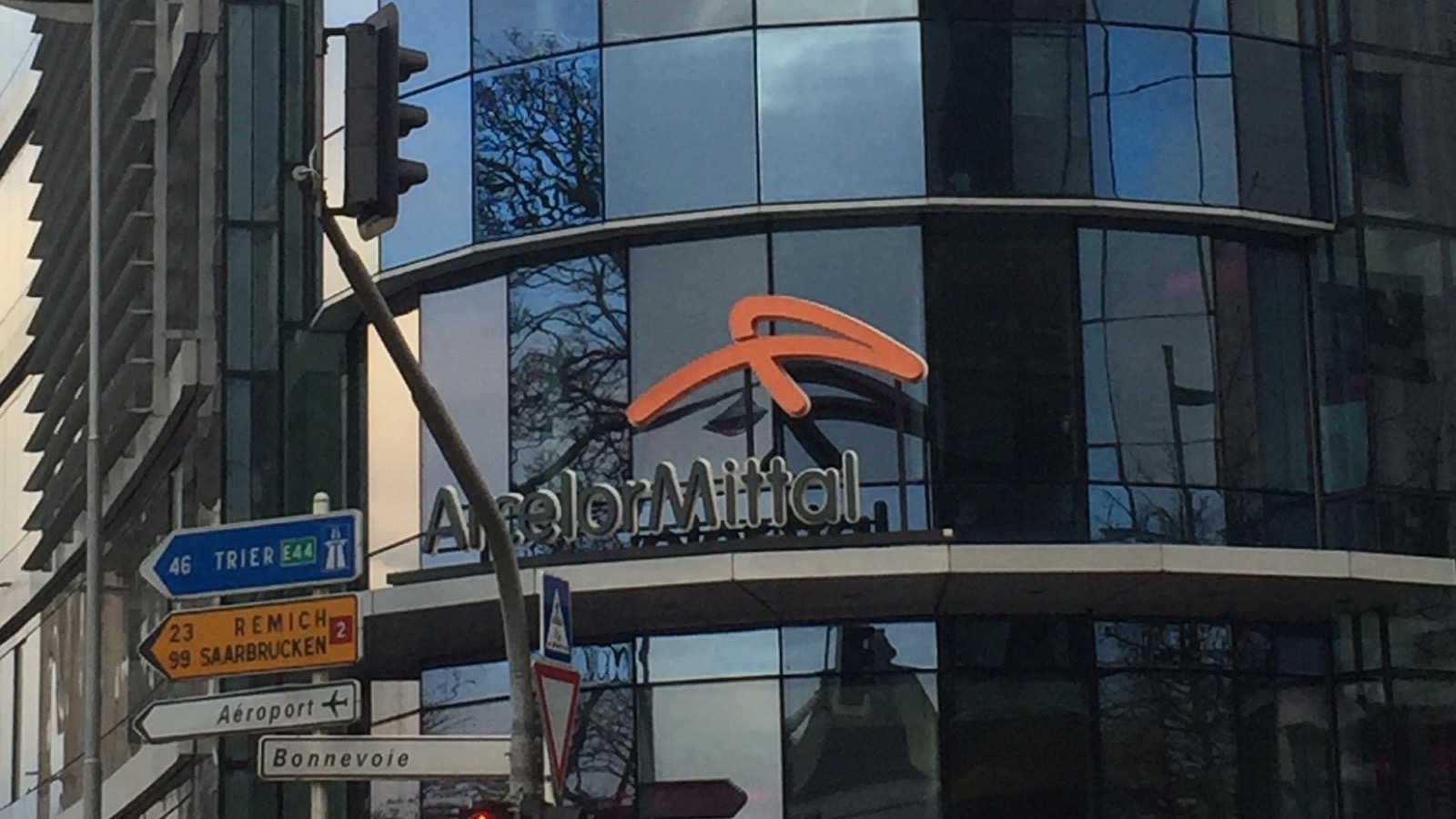 ルクセンブルク市にある ArcelorMittal のグローバル本社。ガラス張りの角地ビルの正面エントランス上に、オレンジ色のロゴマークと「ArcelorMittal」の社名が掲げられている。
