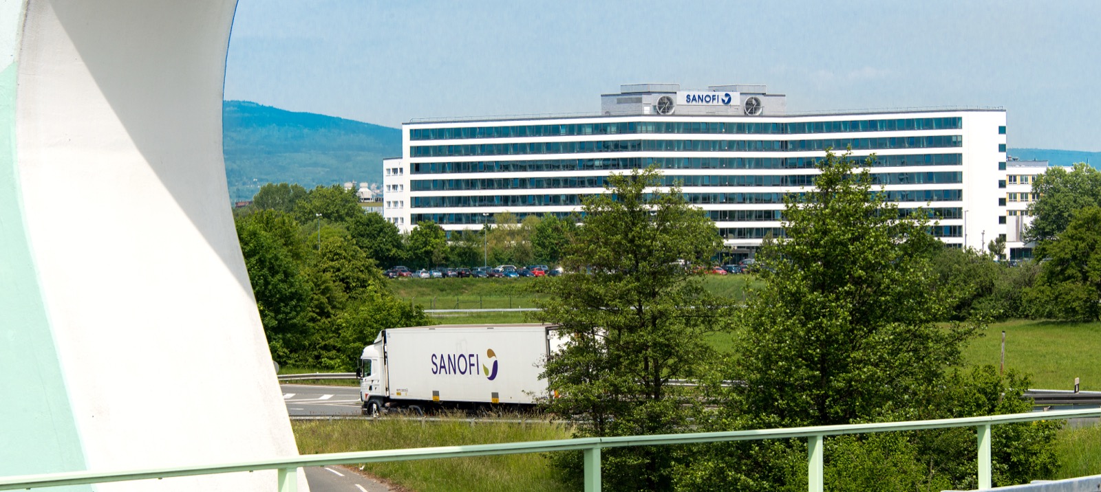 ドイツ・フランクフルトのインダストリーパーク・ヘヒスト内にあるSanofiのドイツ本社。屋上に「SANOFI」ロゴを掲げた白い本社棟と、手前を走るSanofiブランドのトラックが写る。
